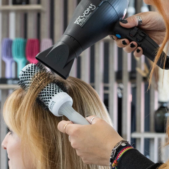 Person using a hair dryer and brush on another person's hair in a salon setting.